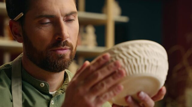 Close up adult focused talented Caucasian salesman potter male man guy businessman artist small business owner holding pottery clay ceramic bowl examining pattern ornament decoration indoors workshop