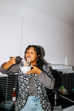 Young female student slurping noodles with eyes closed sitting on kitchen counter in college dorm