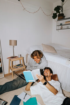 Happy young man showing book to male friend sitting by bed in college dorm