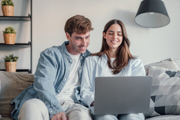 Happy millennial couple sit relax on couch in living room watching video on laptop together, smiling young husband and wife rest on sofa at home browsing Internet using modern computer device.