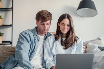 Obraz premium Happy millennial couple sit relax on couch in living room watching video on laptop together, smiling young husband and wife rest on sofa at home browsing Internet using modern computer device.