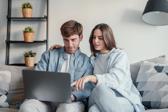 Happy millennial couple sit relax on couch in living room watching video on laptop together, smiling young husband and wife rest on sofa at home browsing Internet using modern computer device. - Powered by Adobe