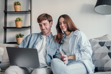 Obraz premium Happy millennial couple smiling looking at laptop screen making video call, young man and woman customers using computer applications for online shopping or booking sitting on sofa embracing.
