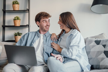 Fototapeta premium Happy millennial couple smiling looking at laptop screen making video call, young man and woman customers using computer applications for online shopping or booking sitting on sofa embracing.