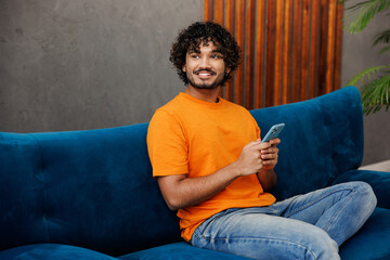 Young smiling Indian man wear orange casual clothes sits on blue sofa couch hold use mobile cell...