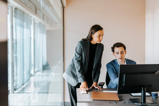 Businesswoman leaning on desk by male colleague using computer while sitting at office