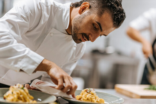 Young male chef cleaning plate after plating pasta at table in kitchen