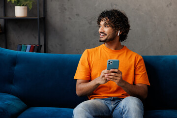 Young Indian man wear casual clothes sits on blue sofa couch listen music in earphones use mobile...