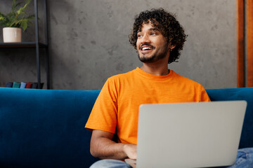 Young happy IT Indian man wear casual clothes sit on blue sofa couch hold use work on laptop pc...