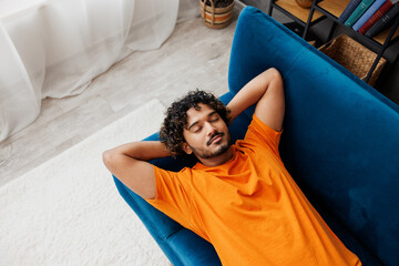 Top view young calm Indian man he wear orange casual clothes lay down on blue sofa couch sleep take...