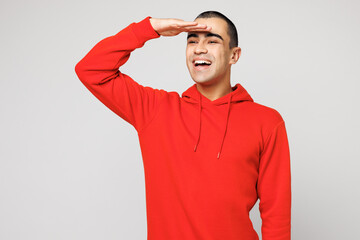 Young smiling happy fun middle eastern man he wears red hoody casual clothes hold hand at forehead look far away distance isolated on plain solid white background studio portrait. Lifestyle concept.