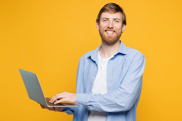 Young smiling smart happy IT man wear blue shirt t-shirt casual clothes hold use work on laptop pc computer chat online isolated on plain yellow orange background studio portrait. Lifestyle concept.