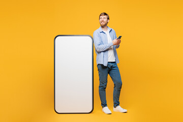 Full body young smiling man wear blue shirt casual clothes stand near big huge blank screen mobile cell phone with area using smartphone isolated on plain yellow orange background. Lifestyle concept.