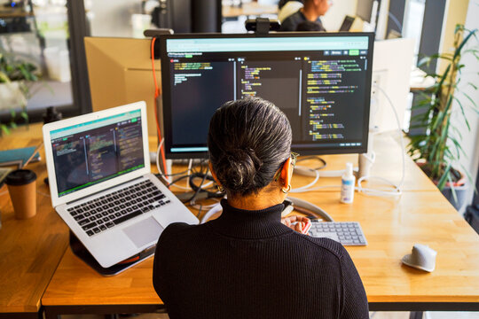 High angle rear view of female programmer coding on desktop PC and laptop while sitting at desk in office