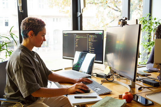 Focused non-binary IT professional working on desktop PC at desk in startup office