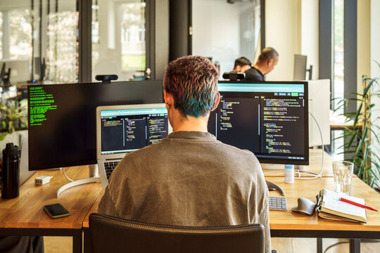 Rear view of non-binary coding professional working on desktop PC and laptop in office