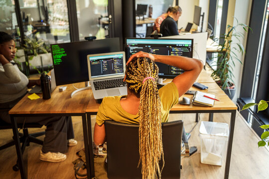 High angle rear view of braided hair female coding professional sitting near desktop PC and laptop in tech hub