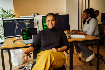 Portrait of confident businesswoman sitting with legs crossed at knee near desktop PC in tech startup office