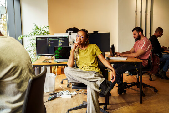 Portrait of smiling female programmer sitting on chair in front of desktop PC at tech startup office