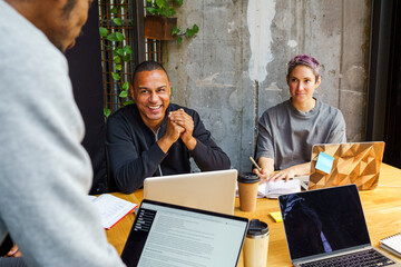 Male business professional sitting next to non-binary colleague with laptop in meeting at office