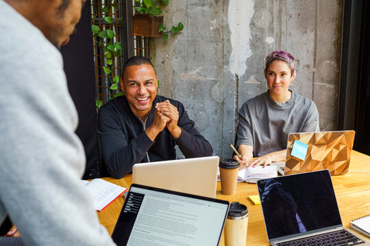 Male business professional sitting next to non-binary colleague with laptop in meeting at office