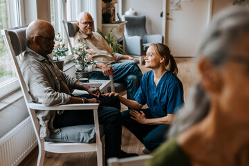 Smiling nurse talking with elderly man sitting on chair at care home