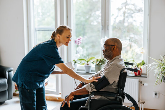 Female medical expert fastening blood pressure gauge on senior man's arm sitting in wheelchair at nursing home