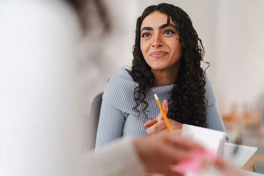 A young woman with curly hair wearing a light blue sweater is engaged in discussion, holding a yellow pencil in her hand while speaking in a bright and modern workspace.