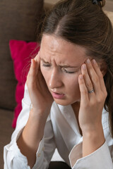 Stressed young woman in a white shirt suffering from a migraine, experiencing a throbbing headache,...