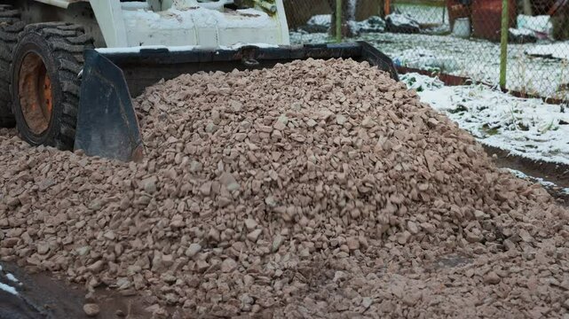 A small construction vehicle distributes crushed stones on a pathway in a winter setting. Snow patches, trees, and a chain link fence are visible.