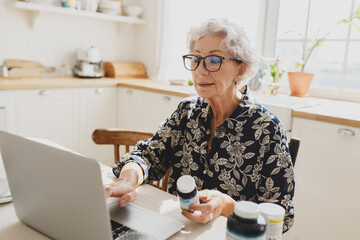 Side view of smart grandmother in glasses sitting at kitchen table and ordering food supplements online on market place or pharmacy holding bottle of vitamins and typing on laptop its name
