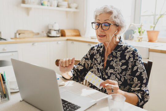 Senior pretty grandmother with gray hair in glasses consulting with her doctor online using earphones, laptop and video chat application, holding blister with pills. Telemedicine concept