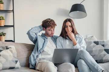 Millennial couple annoyed by stuck laptop or online news sitting together on sofa, frustrated young...