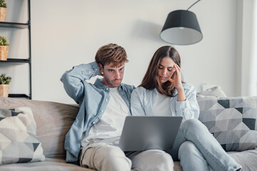 Millennial couple annoyed by stuck laptop or online news sitting together on sofa, frustrated young...