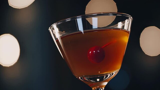 Glass of Manhattan cocktail with a deep amber hue, garnished with a maraschino cherry, served in a martini glass against a moody bokeh-lit background