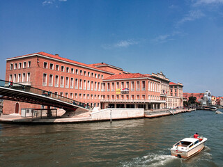 A View of Santa Lucia Station in Venice. The station building, with its distinctive red brick facade, is a prominent landmark along the canal. A boat passes by, adding movement and life to the scene.