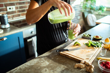 Senior woman pouring green smoothie into glass in kitchen with fresh ingredients on countertop