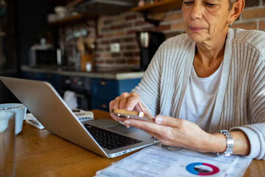 Senior woman using smartphone for online banking next to laptop at home