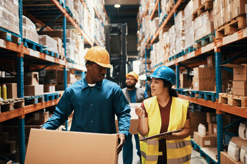 Warehouse workers discussing logistics while carrying boxes and using clipboard