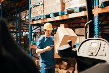 Smiling male worker using smartphone and carrying box in large warehouse