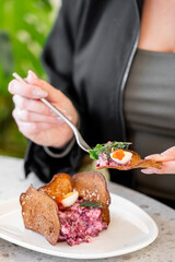 A close-up shot of a person's hand holding a spoonful of vibrant beetroot salad, topped with herbs and small eggs, served on a crispy bread base. The background features lush greenery.