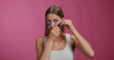 A woman applies patches to her lower eyelids. She smiles at the camera while standing on a pink background in the studio