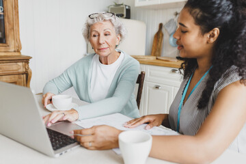 Side view of young african american female social worker teaching elderly caucasian lady to use laptop and improving her skills, sitting together at kitchen table and having tea or coffee