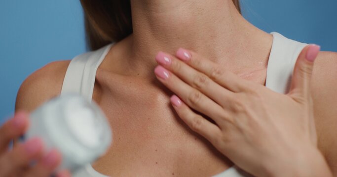 A woman applies a cosmetic cream to her neck while standing on a studio background. Close-up of a body part, an unrecognizable person. She holds the bottle in her hand