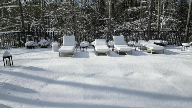 aerial view of snow covered lounge chairs next to closed shuttered swimming pool in winter (back yard patio loungers outdoor furniture) spa bizzard snowy snowfall frozen home countryside drone footage