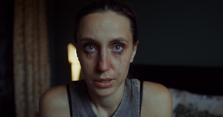 A woman is crying sitting on a bed at home. Her mascara was leaking. She's looking at the camera