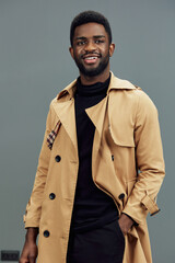 Stylish young African man wearing a beige trench coat, smiling confidently against a muted gray background, perfect for fashion and lifestyle concepts