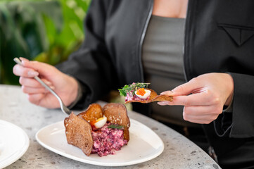 A person holds a fork and a bite of gourmet salad topped with herbs and sauce, served on a white plate. The setting features lush greenery, enhancing the fresh and vibrant atmosphere of the dish.