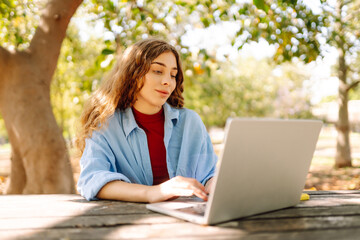 Attractive woman working or studying at the laptop as sitting in city park. Freelancer working in the park. Online course