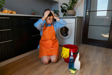 Woman holding her head in exhaustion from housework. Cleaning supplies and a bucket with wet rags...